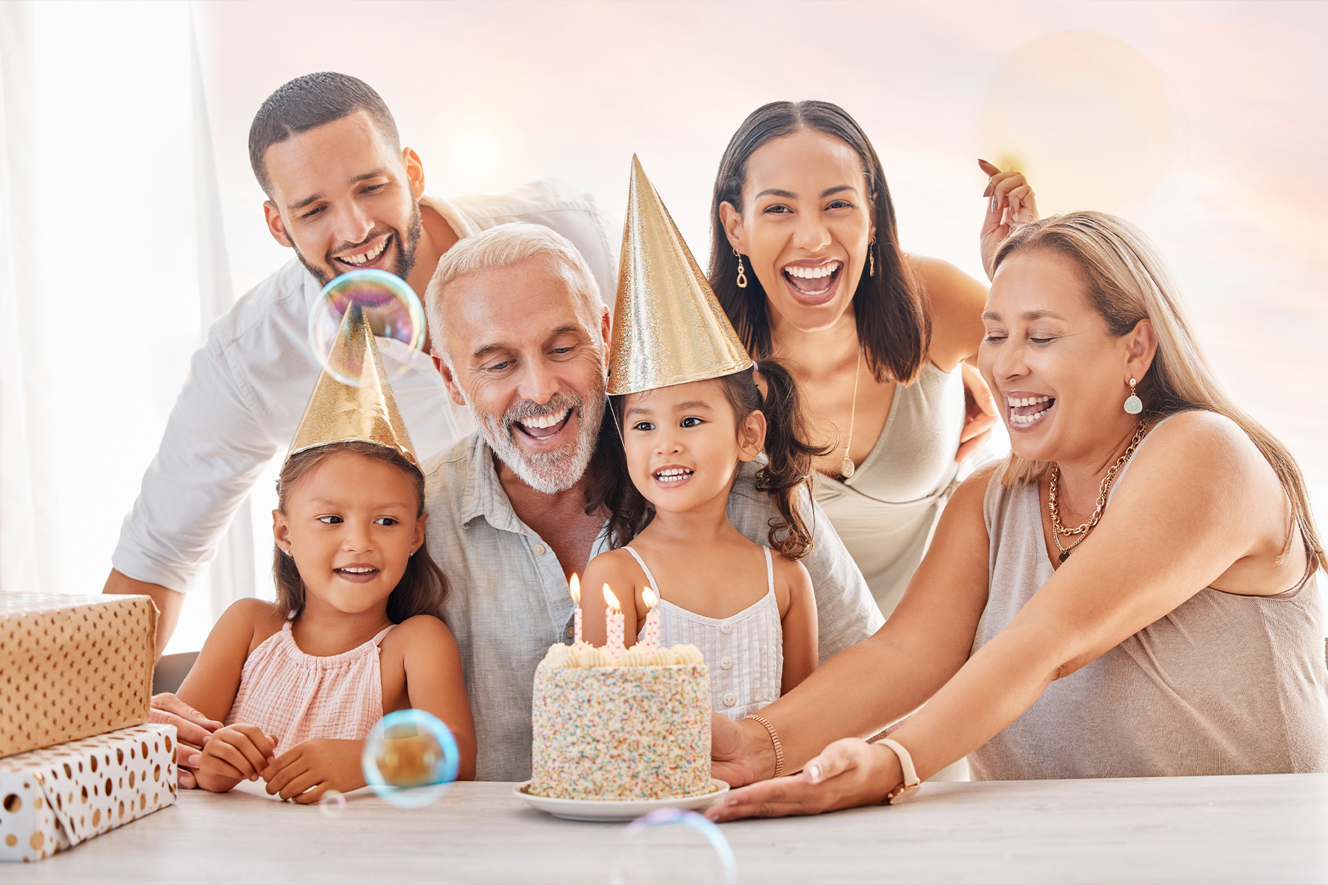 A multigenerational family with party hats laughing and celebrating around a birthday cake with lit candles