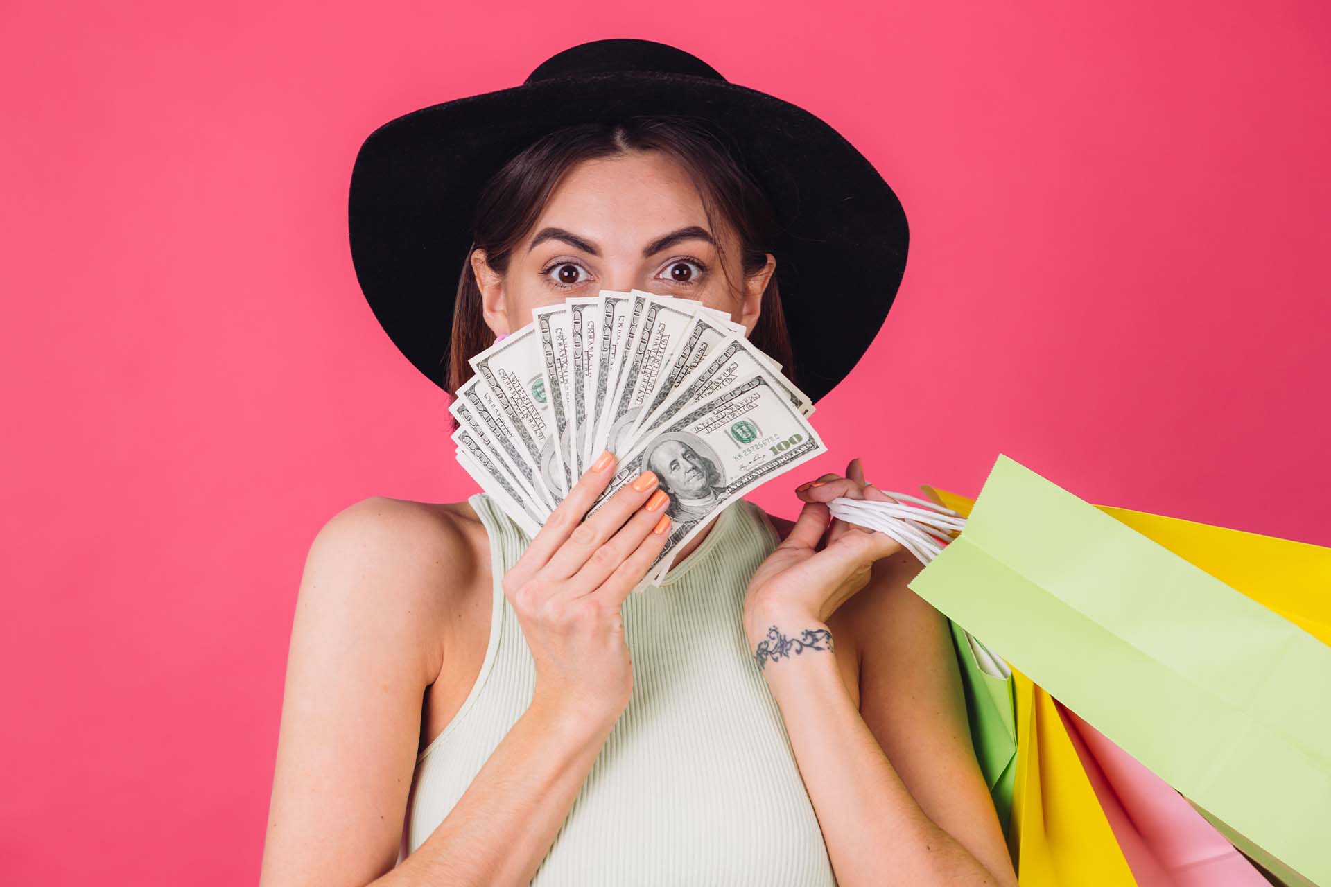A woman in a white tank top and black hat, holding a fan of cash and colorful shopping bags, symbolizing impulsive spending